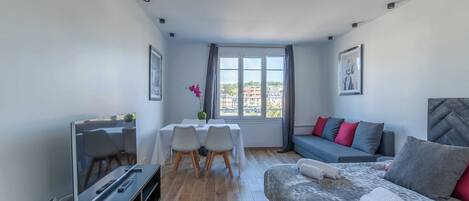 A modern hotel room with a bed, a sofa, a dining table with chairs, a TV, and a window with curtains.