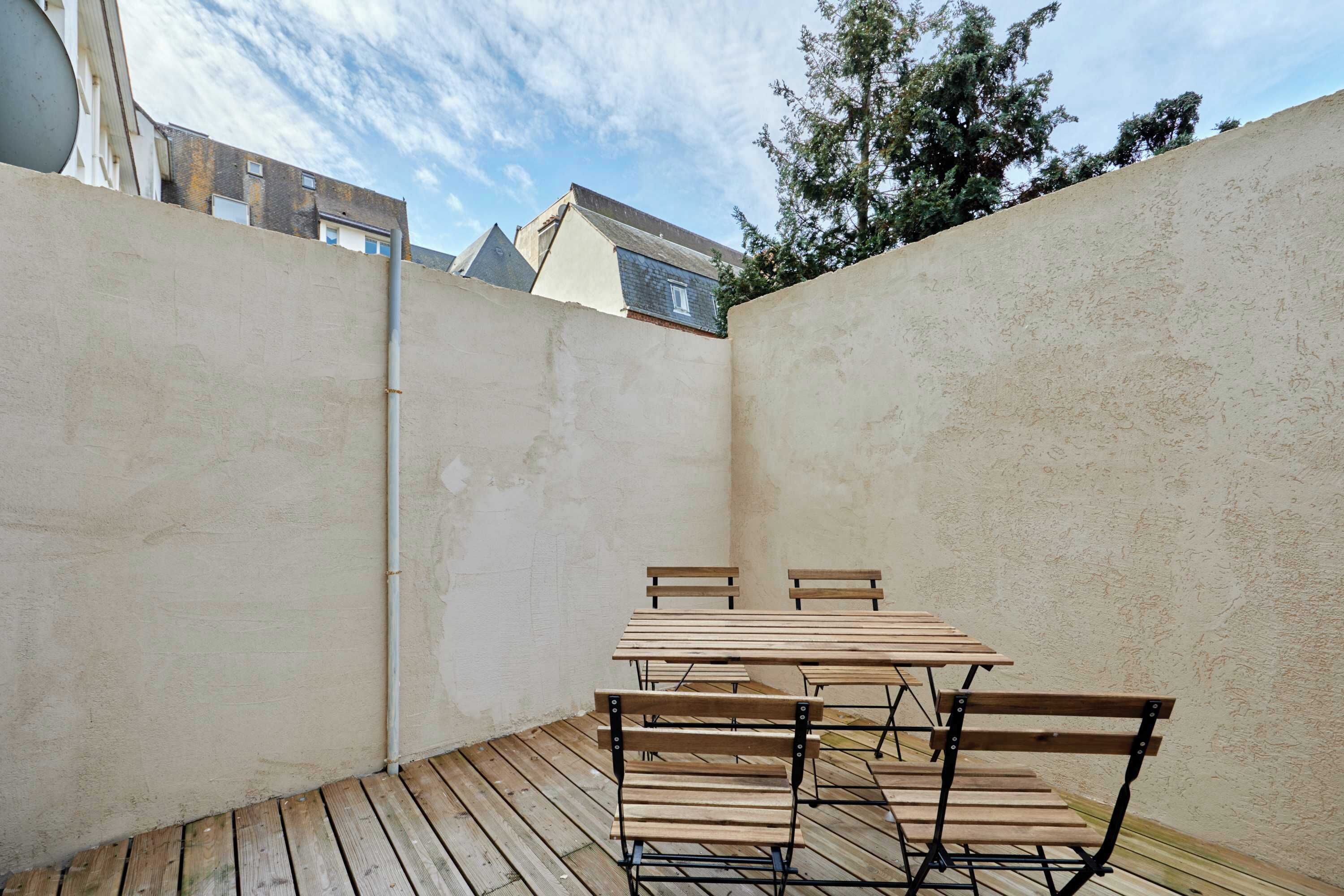 A patio area with a wooden table and chairs, surrounded by high walls and a wooden deck.