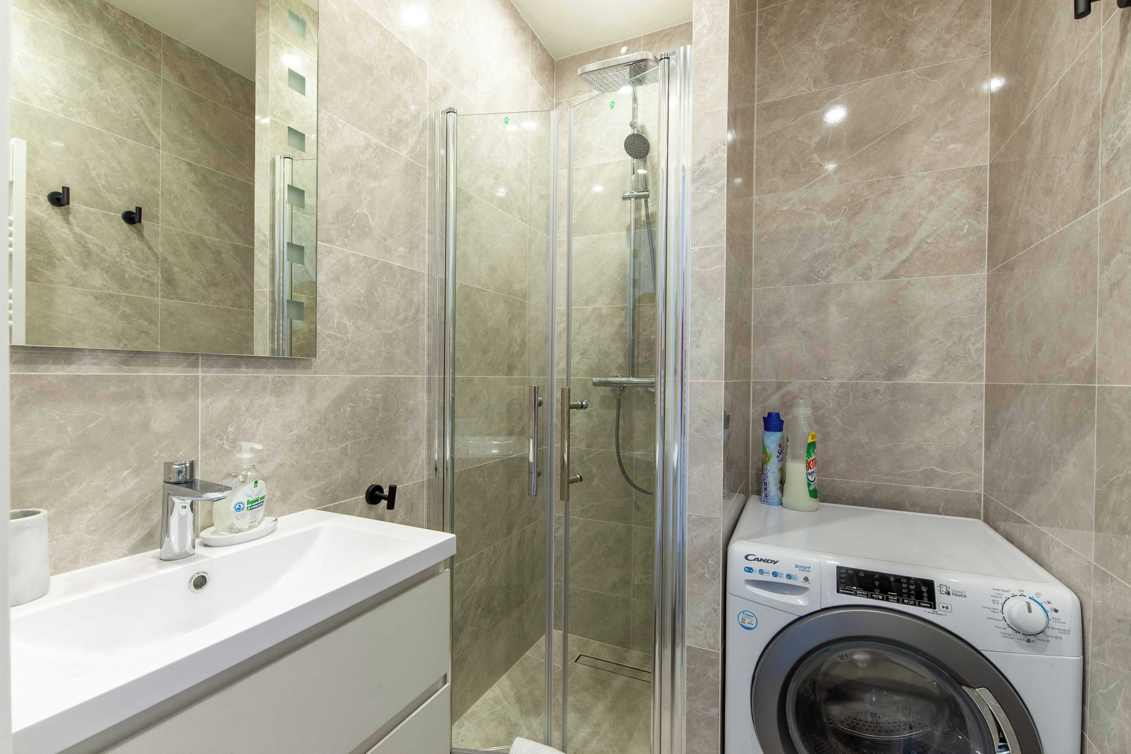 Un baño moderno con ducha envuelta en cristal, un lavamanos blanco con dispensador de jabón y una lavadora de carga frontal.