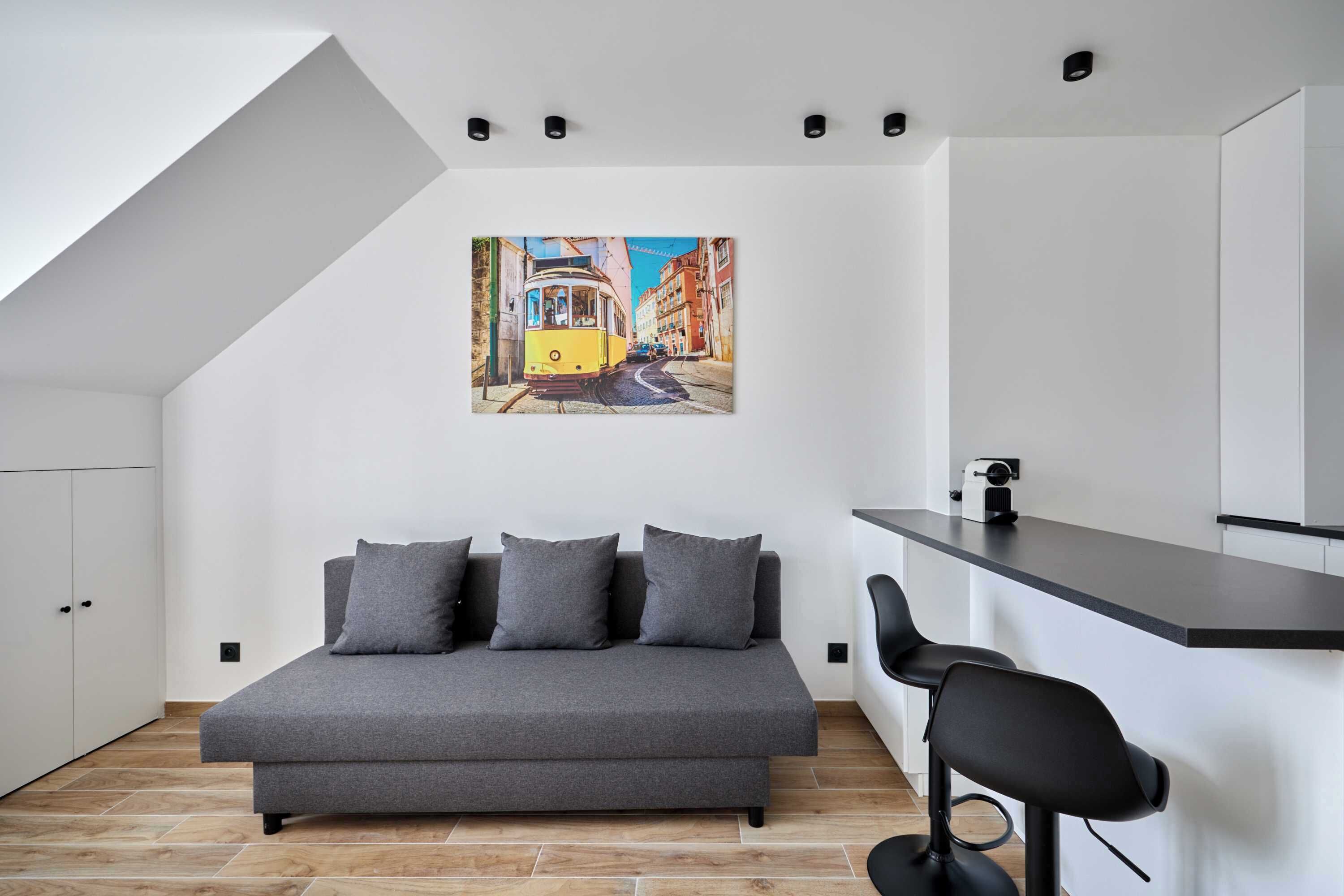 Quarto moderno com sofá cinzento, um pequeno bar e um quadro de um elétrico na parede.