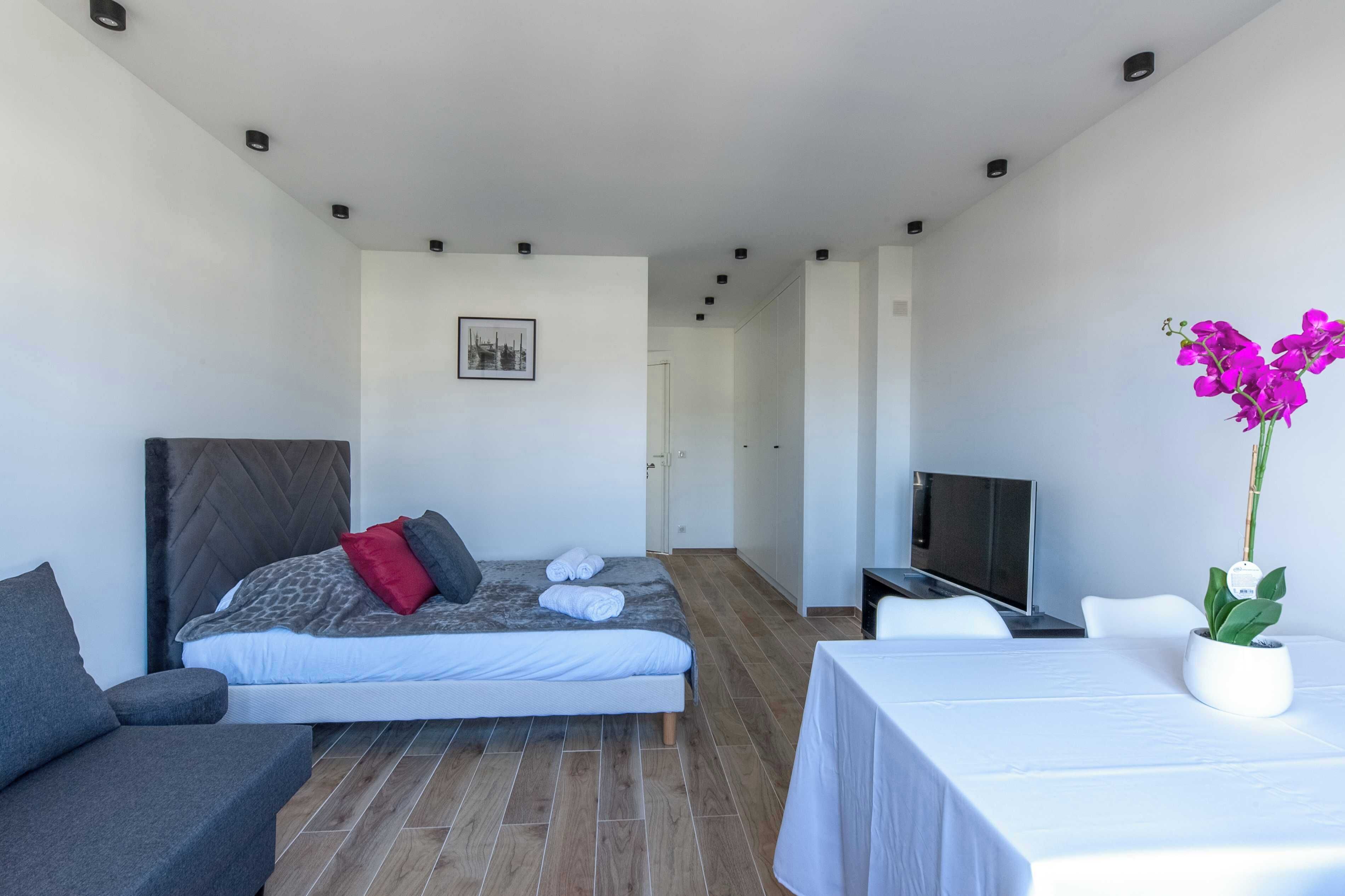 A modern hotel room with a bed, a TV, a sofa, and a table with a plant.