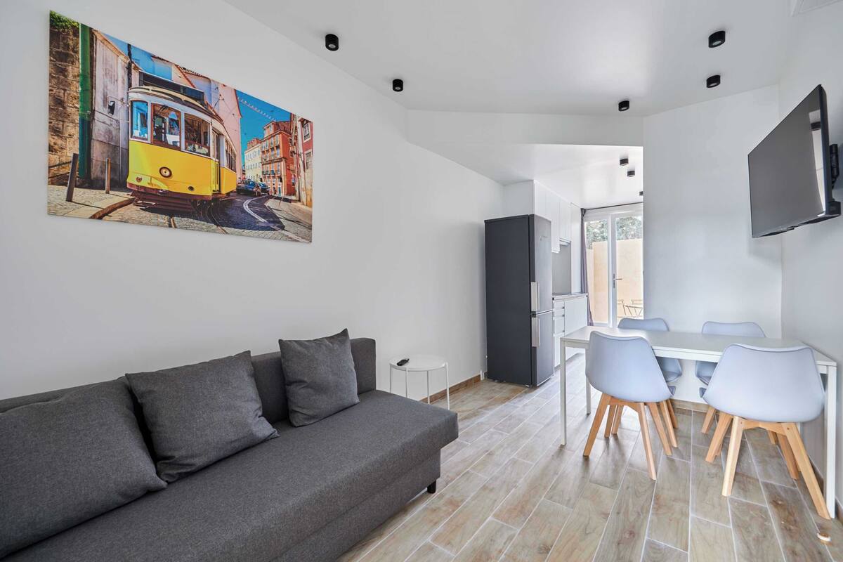 Un salon moderne avec un canapé gris, une table à manger avec des chaises, un téléviseur fixé au mur et un tableau représentant un tramway accroché au mur.