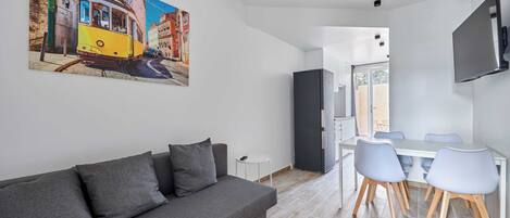 A modern living room with a grey sofa, a dining table with chairs, a TV mounted on the wall, and a painting of a tram on the wall.