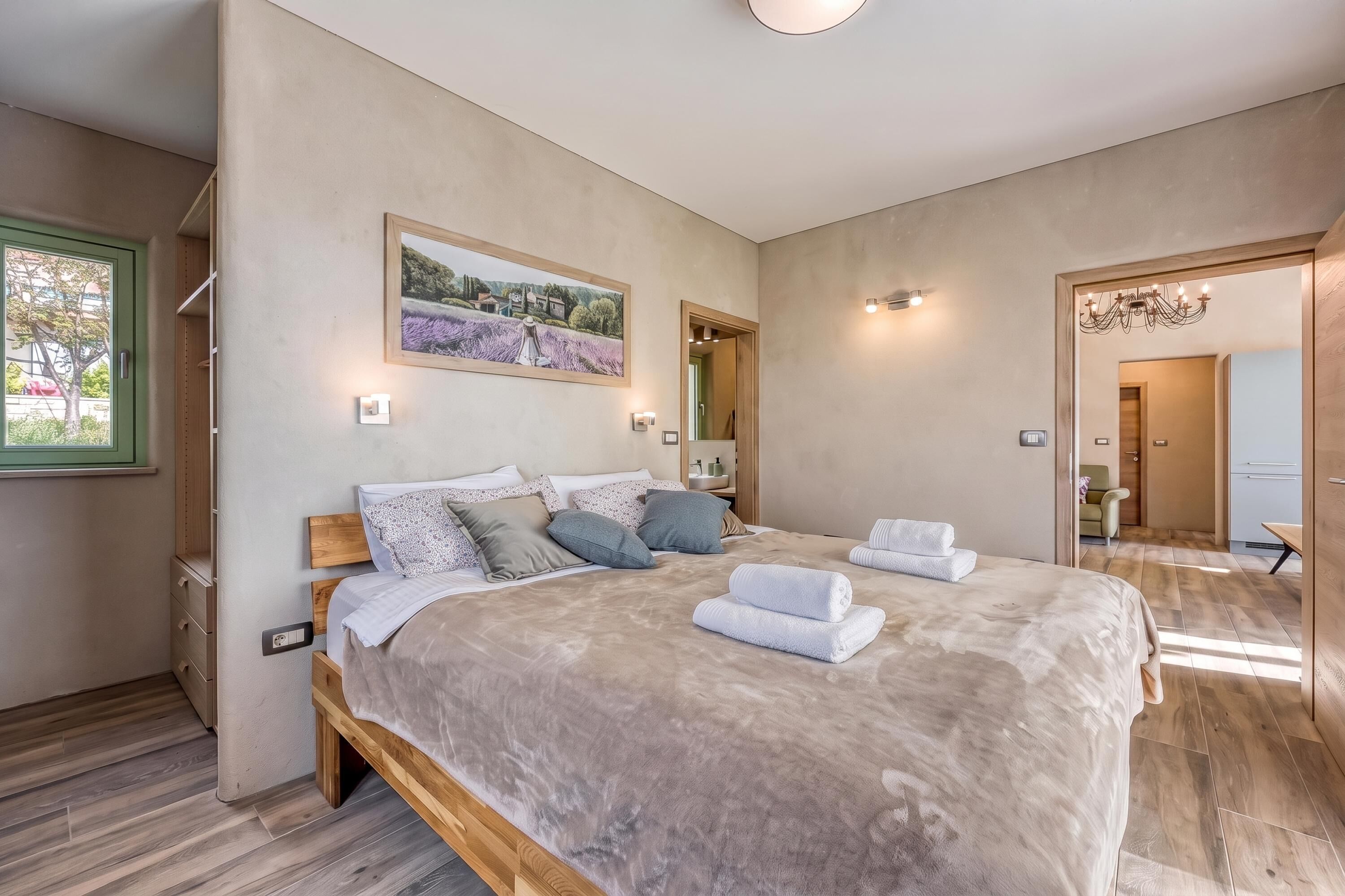 A bedroom with a large bed, a painting on the wall, and a view of the outside.