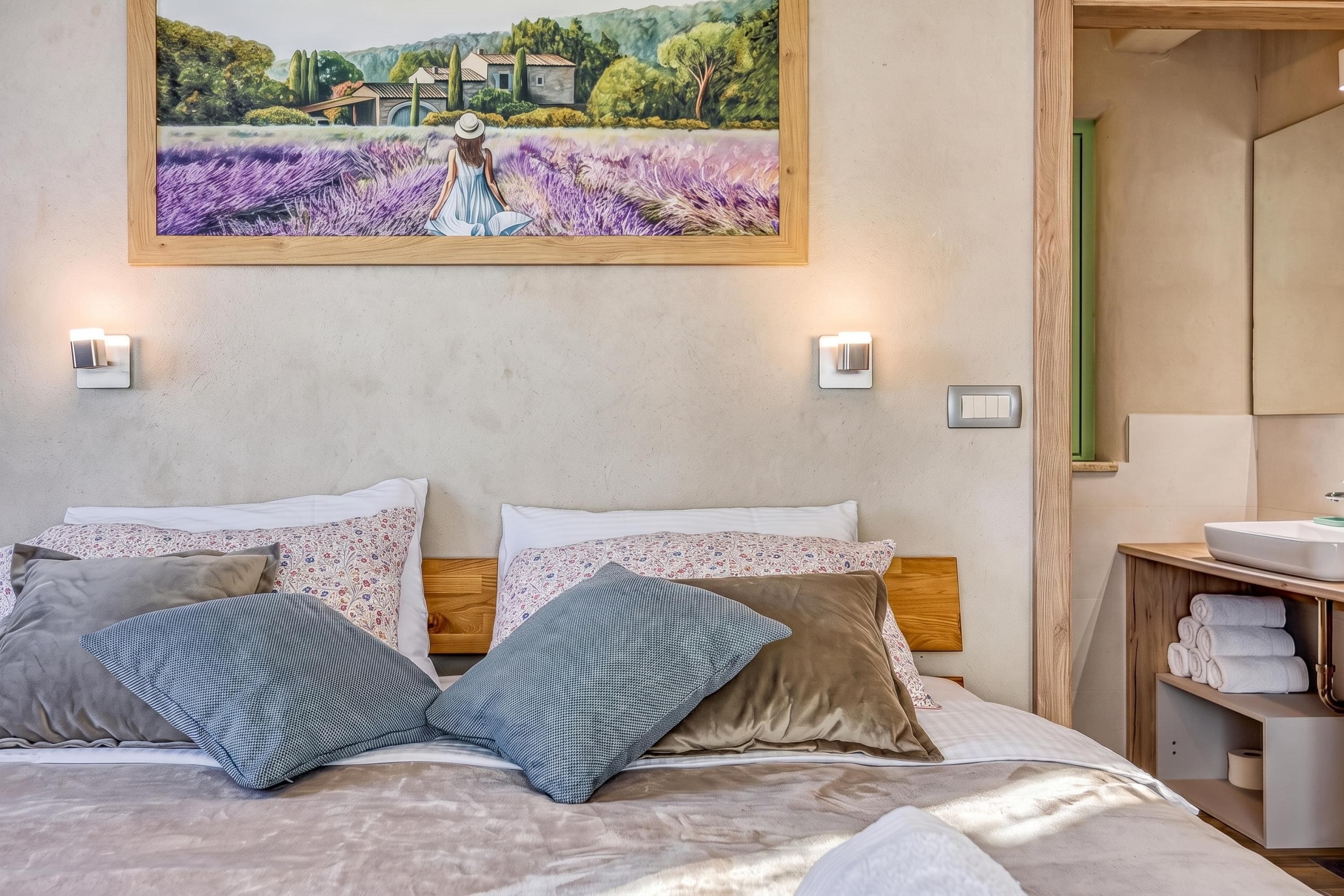 A bedroom with a bed, pillows, a painting, a bedside table, and a bathroom visible through an open door.