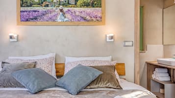 Un dormitorio con cama, almohadas, un cuadro, una mesita de noche y un baño visible a través de una puerta abierta.