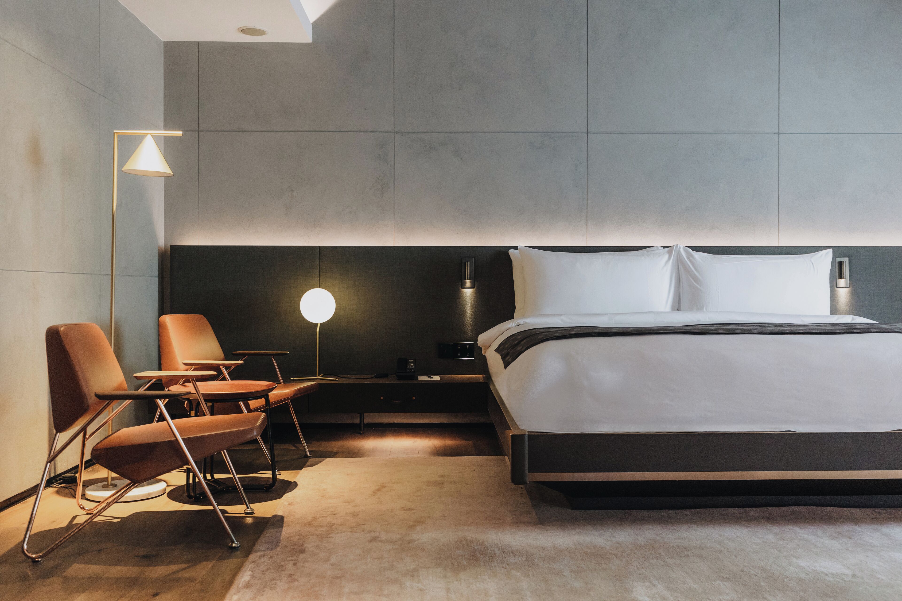A modern hotel room with a large bed, a bedside table, a lamp, and two chairs.