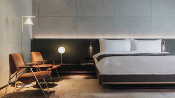 A modern hotel room with a large bed, a bedside table, a lamp, and two chairs.