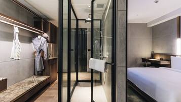 A modern hotel room with a glass-enclosed shower, a bed, a desk, and a seating area.
