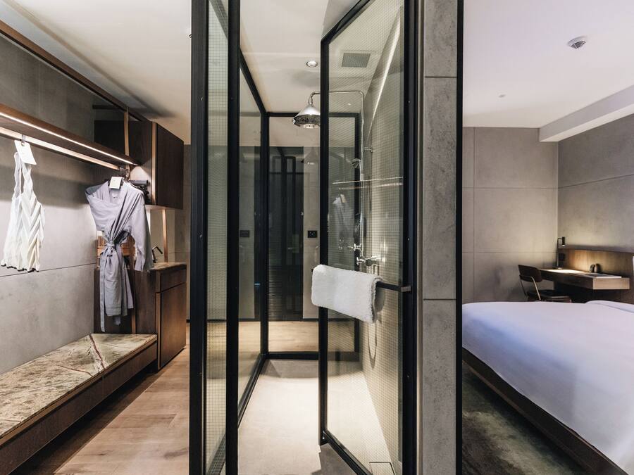 A modern hotel room with a glass-enclosed shower, a bed, a desk, and a seating area.