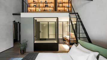 A modern hotel room with a large bed, a small bathroom, a staircase leading to a mezzanine with shelves, and a view of the city.