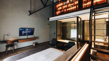 A modern hotel room with a bed, a desk with a computer, a TV, and a seating area with a small table and chairs.