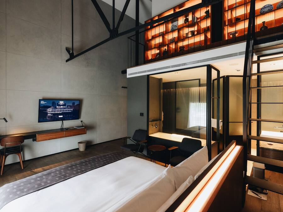 A modern hotel room with a bed, a desk with a computer, a TV, and a seating area with a small table and chairs.