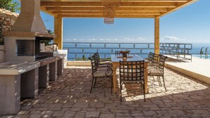 Een terras buiten met een stenen vloer, een houten pergola, een barbecue en een tafel met stoelen.