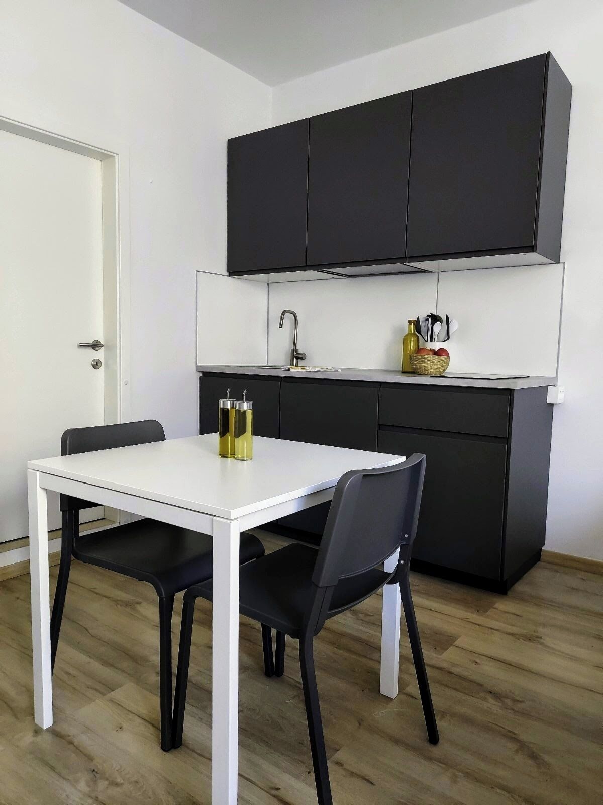 Uma cozinha compacta com uma mesa branca e cadeiras pretas, um armário preto acima do balcão e uma pia pequena.