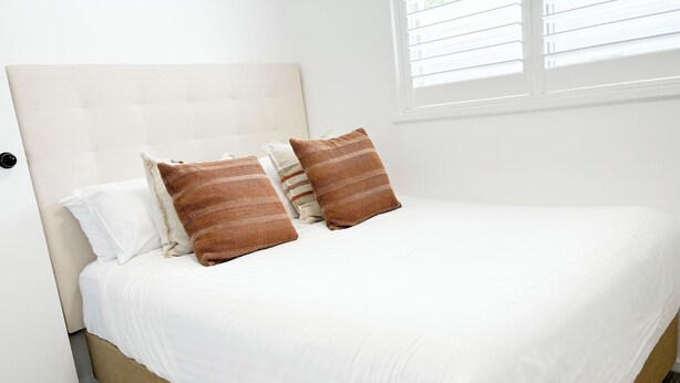 A neatly made bed with white bedding and two patterned pillows, a beige headboard, and a window with blinds. - Haven in the heart of Turvey Park! (Wagga Wagga)