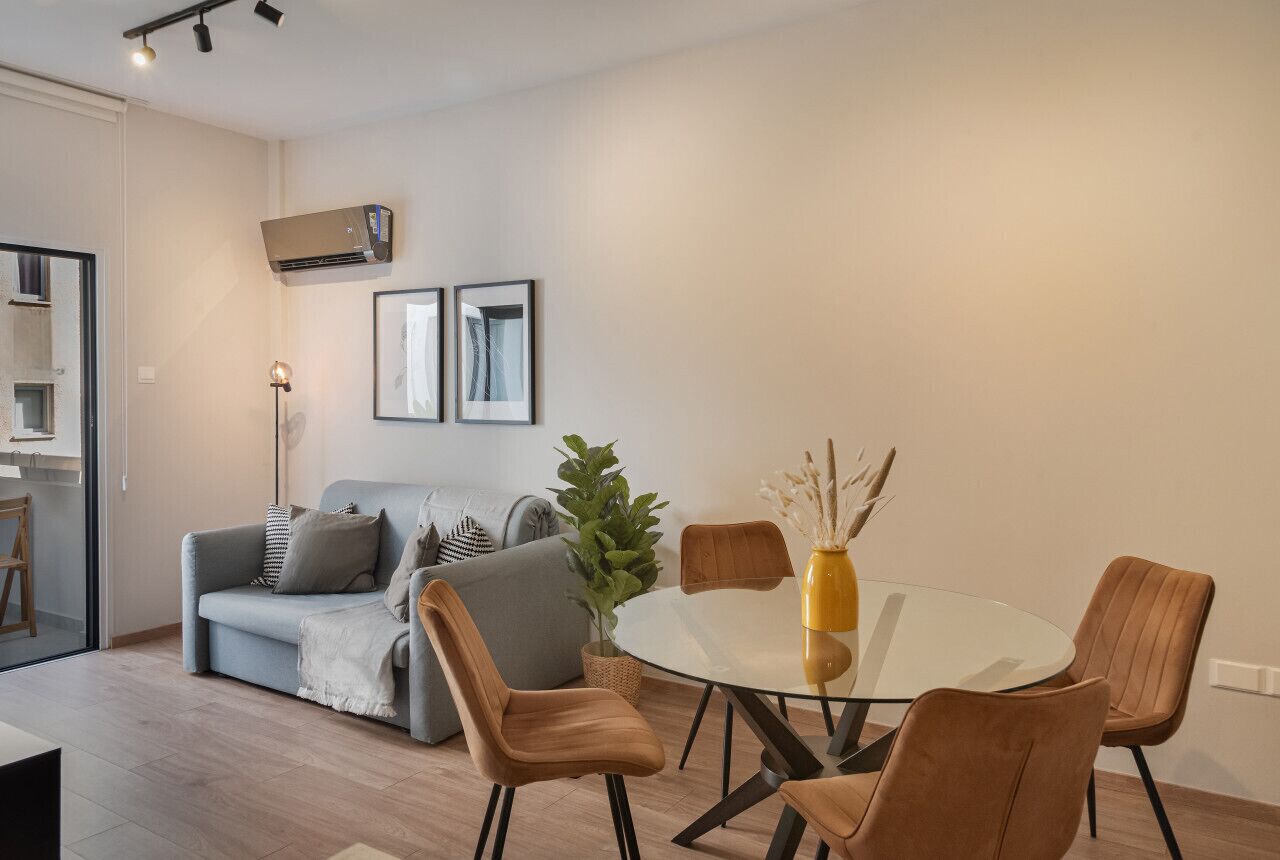 A modern living room with a glass dining table, brown chairs, a grey sofa, and a potted plant.