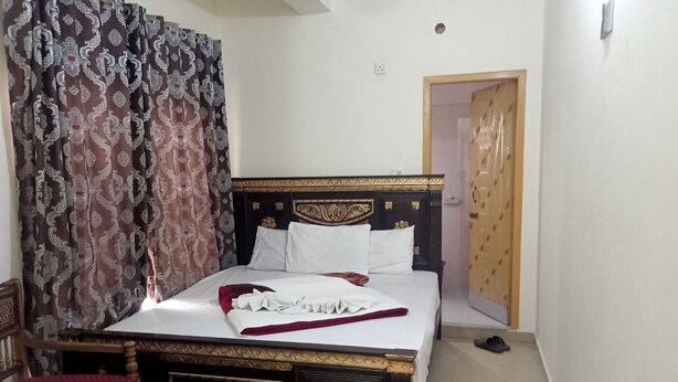 A neatly made bed with white linens, a wooden headboard, and a red throw blanket. There is a small table with a glass top and a plate with a spoon. The room has patterned curtains and a wooden door. - Nest Hotel (Rawalpindi)