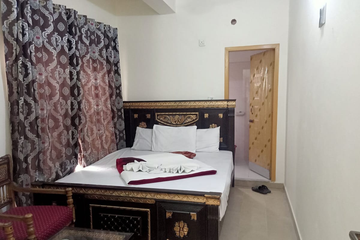 A neatly made bed with white linens, a wooden headboard, and a red throw blanket. There is a small table with a glass top and a plate with a spoon. The room has patterned curtains and a wooden door.