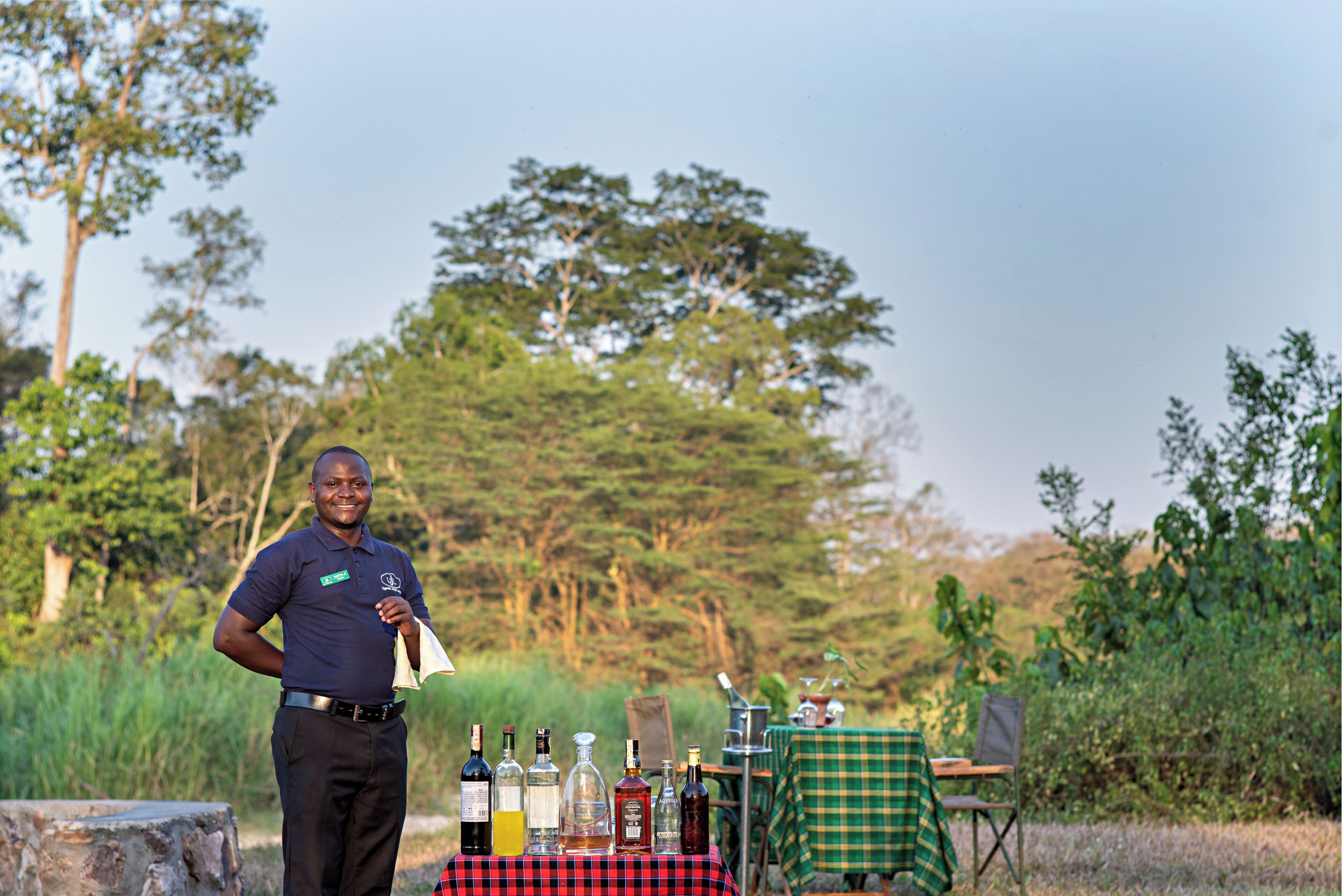 A person standing in front of a table with various bottles, possibly for a tasting event, in an outdoor setting with trees in the background.