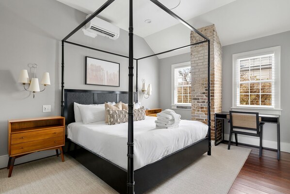 A modern bedroom with a four-poster bed, a wooden nightstand, a desk with a chair, and a large window with blinds. - Thomas Waring House | Unit D | Sleeps 6 (Charleston)