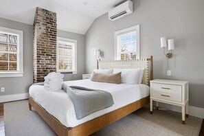 A bedroom with a stone fireplace, a bed with white bedding, a nightstand, and a wall-mounted lamp. - Thomas Waring House | Unit D | Sleeps 6 (Charleston)
