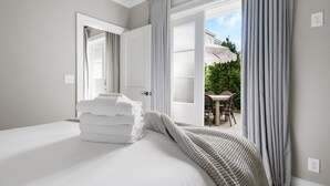 A hotel room with a bed, a stack of towels, and a view of an outdoor patio with a table and chairs. - Thomas Waring House | Unit D | Sleeps 6 (Charleston)