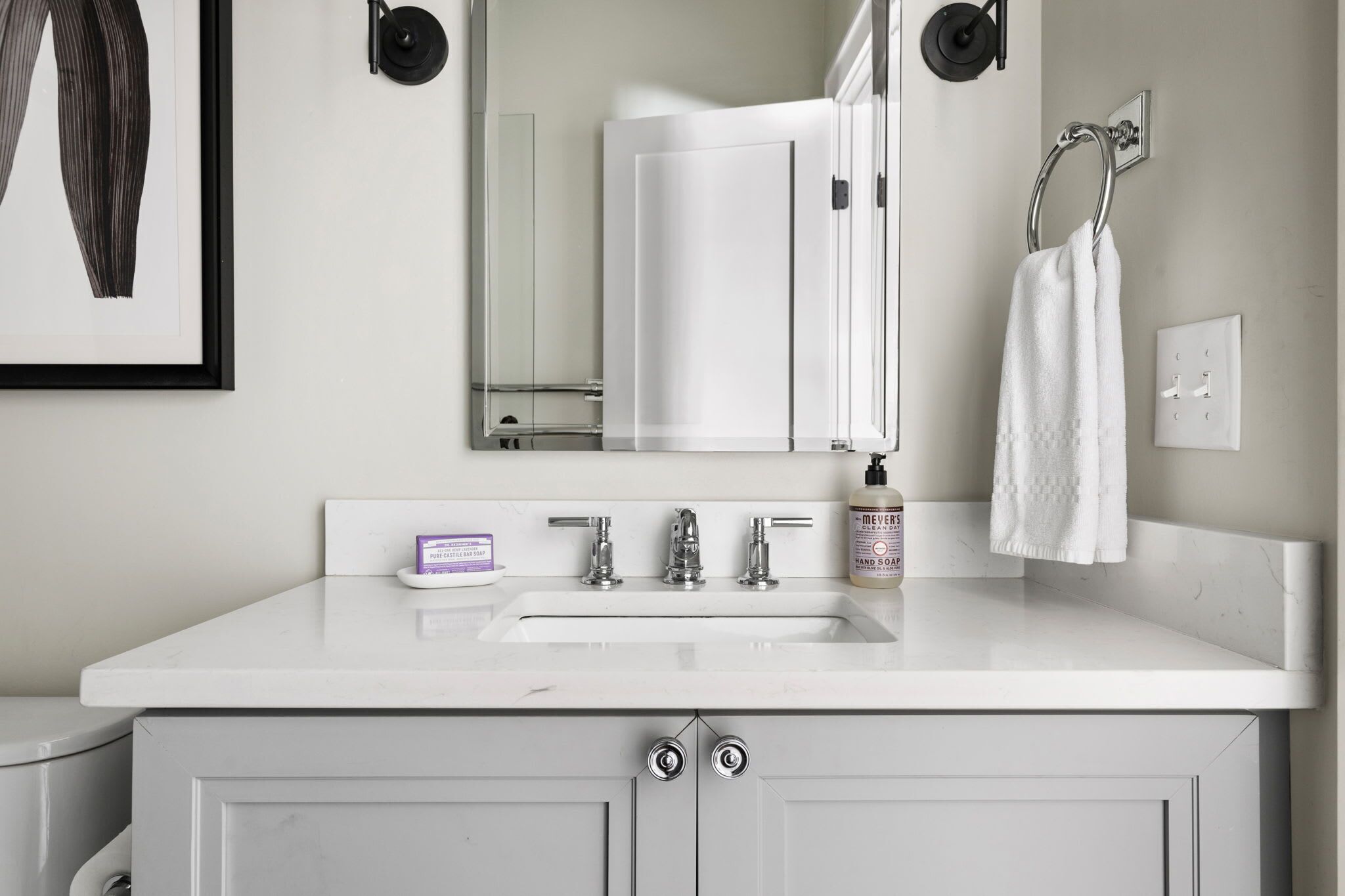 A bathroom with a white sink, two faucets, a mirror, a towel rack, and a bottle of hand soap.