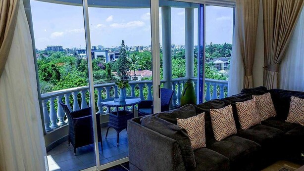 A balcony with a sofa and chairs, a table, and a view of a cityscape and greenery. - Clalem Bay Condos by RevSerene (Mombasa)