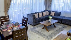 A living room with a sofa, coffee table, and dining area with chairs and a table set for a meal. - Clalem Bay Condos by RevSerene (Mombasa)