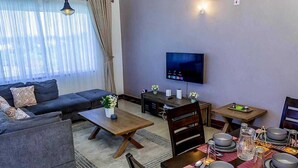 A modern living room with a sofa, coffee table, dining table set for four, a flat-screen TV mounted on the wall, and a large window with sheer curtains. - Clalem Bay Condos by RevSerene (Mombasa)