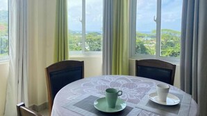 Un coin repas avec une table dressée pour deux personnes, deux chaises et une vue sur un paysage verdoyant grâce à de grandes fenêtres.