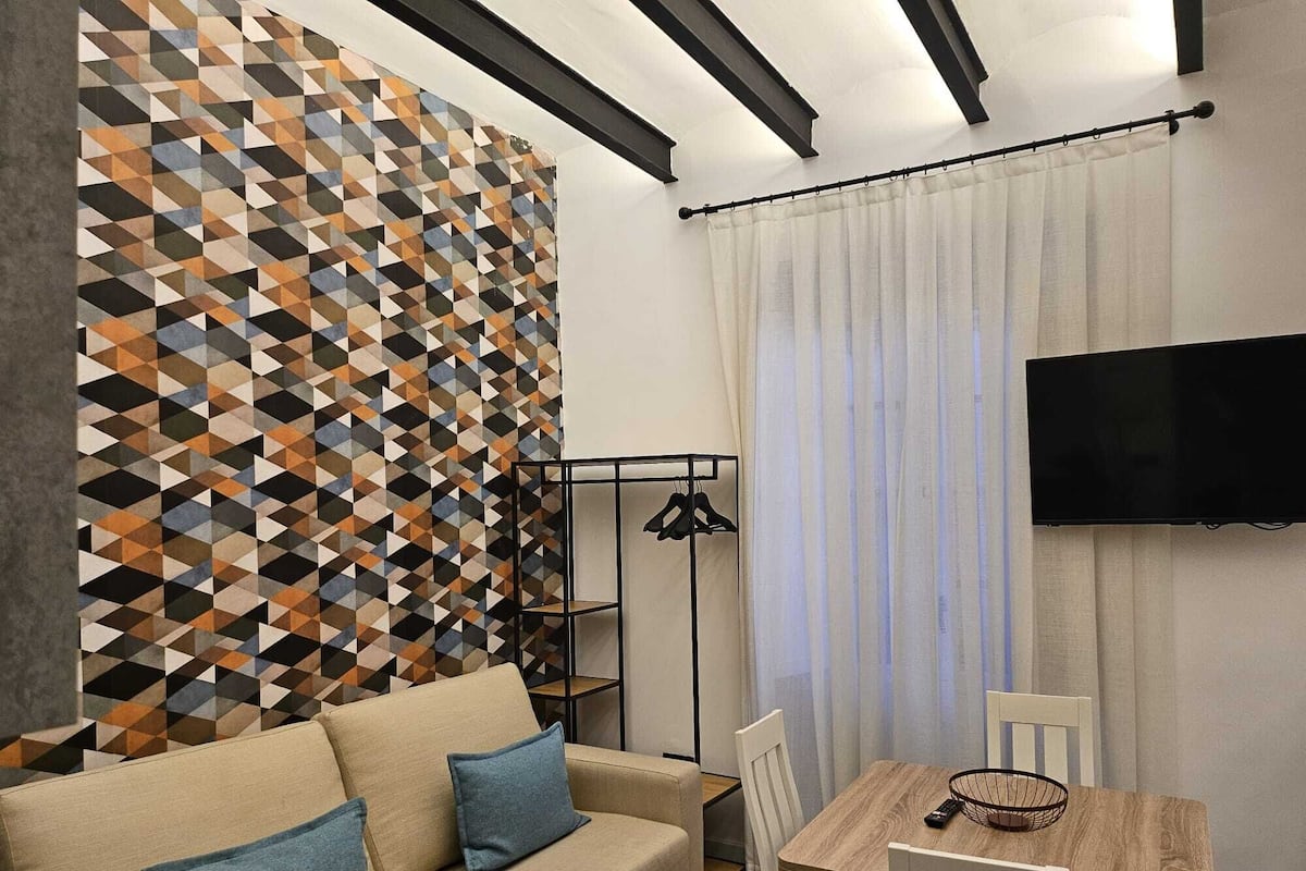 A modern living room with a geometric-patterned wall, a beige sofa with blue cushions, a wooden dining table with chairs, a TV mounted on the wall, and a floor lamp.