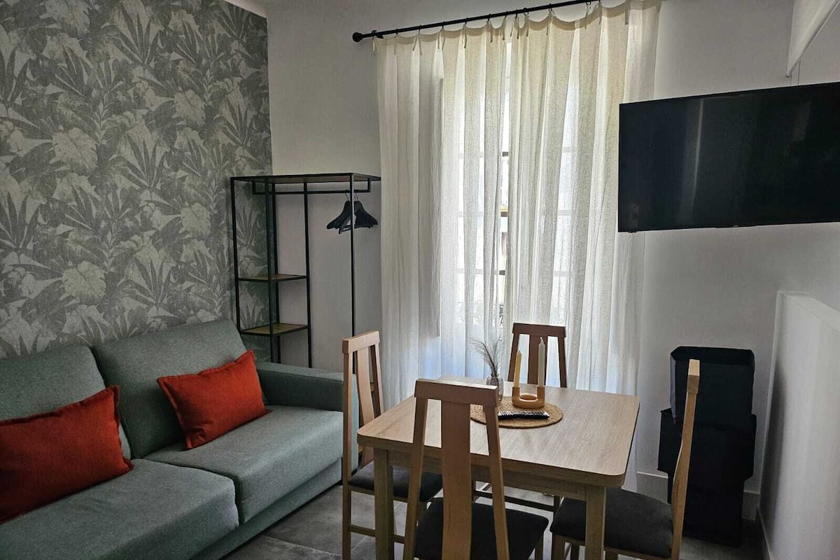 A living room with a grey sofa, red cushions, a wooden table with chairs, a TV mounted on the wall, and a window with sheer curtains.