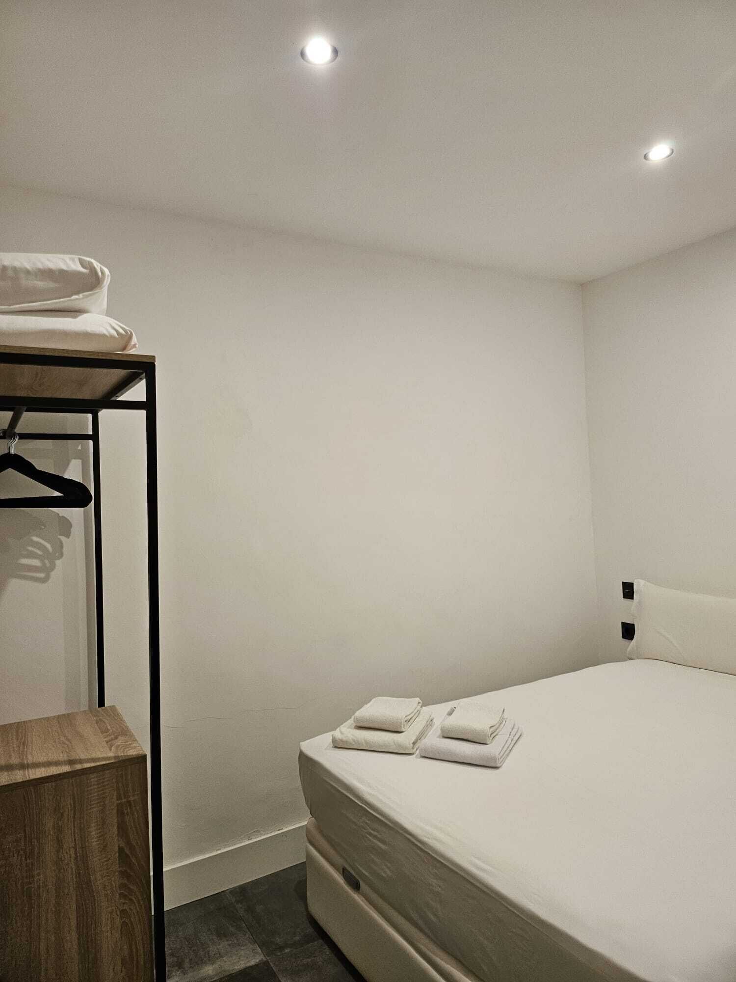 A minimalist hotel room with a bed, a wooden side table, and a wall-mounted shelf with a white towel.