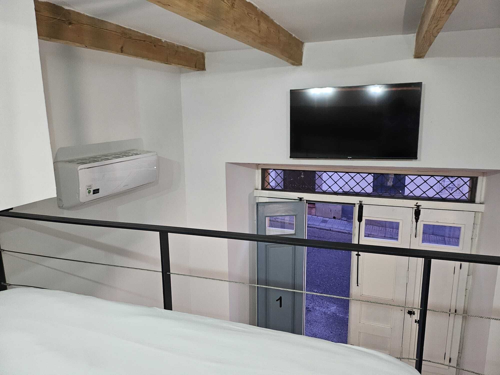 Une chambre dotée d’un lit, d’un téléviseur fixé au mur, d’un appareil de climatisation et d’une fenêtre donnant sur la rue.