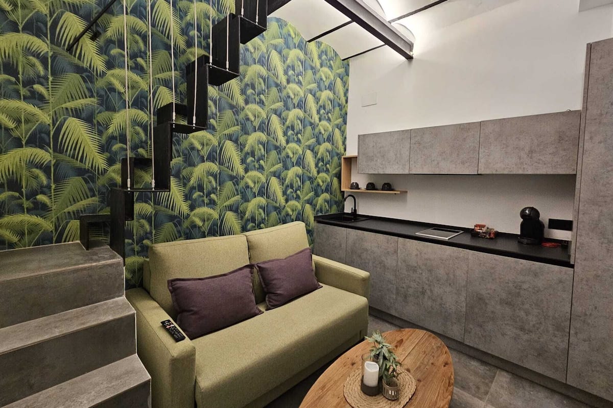 A modern living space with a green sofa, a coffee table, a kitchen area with grey cabinets, and a staircase with a palm leaf wallpaper.