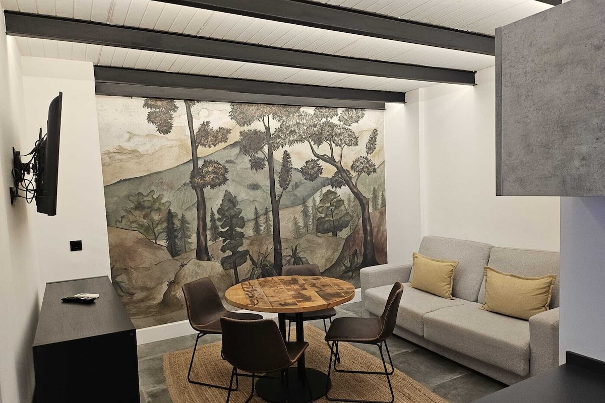 A modern living room with a round wooden table, four chairs, a grey sofa with cushions, and a large mural of a forest scene on the wall.