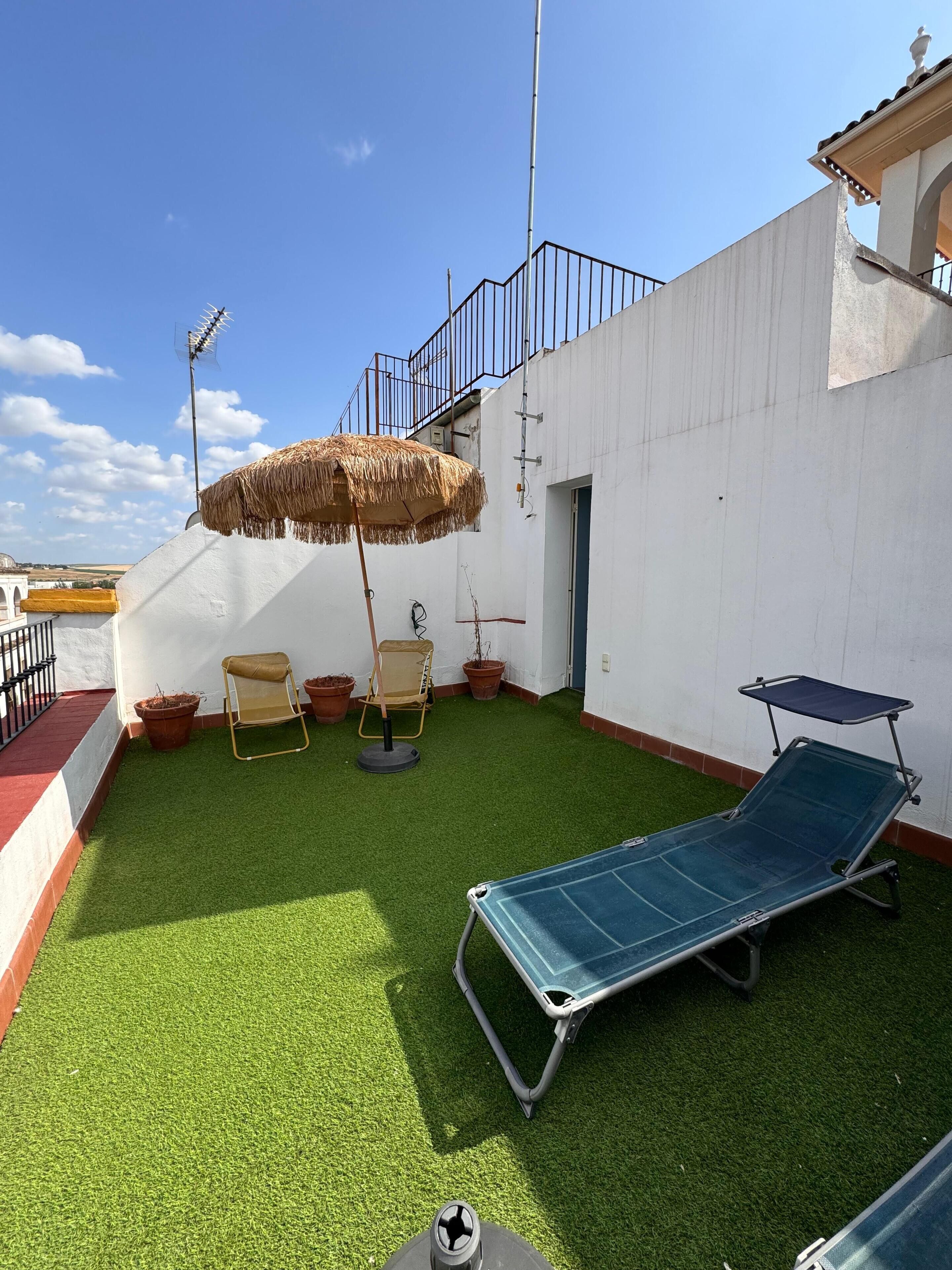 A rooftop terrace with artificial grass, a lounge chair, a sun umbrella, and potted plants.