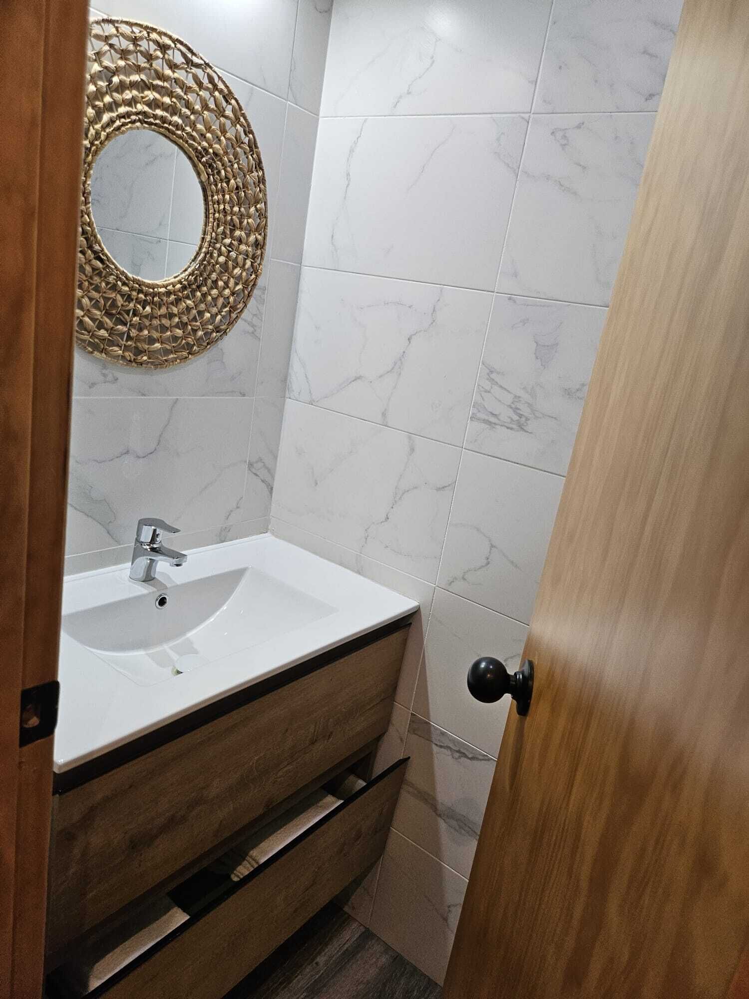 Un baño con lavamanos blanco, tocador de madera, azulejos de mármol y un espejo redondo.