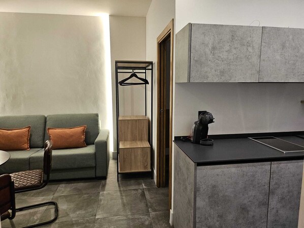 A compact living space with a kitchenette, a sofa with cushions, a chair, and a wooden cabinet. - APARTAMENTOS SAN FERNADO 32 (Córdoba)