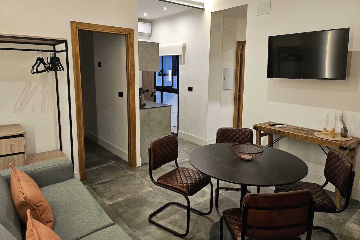 A modern living room with a round dining table, leather chairs, a flat-screen TV, and a built-in kitchen in the background.