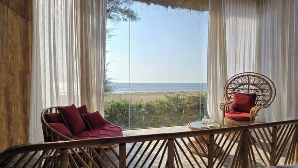 Una habitación con vista a la playa, una silla de mimbre con cojines rojos y una pequeña mesa con un vaso.