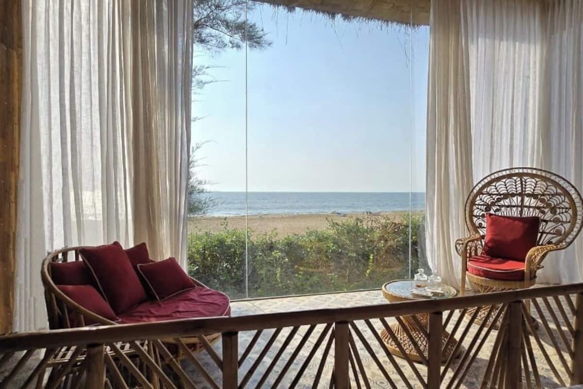 A room with a view of the beach, a wicker chair with red cushions, and a small table with a glass.