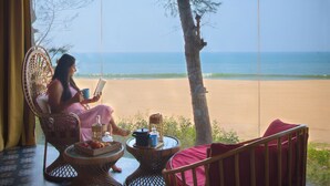 Habitación con un amplio ventanal con vistas a una playa, amueblada con sillas de mimbre y una mesa con una bandeja de frutas.