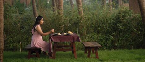 A person sitting at a wooden table outdoors with a grassy area and palm trees in the background.