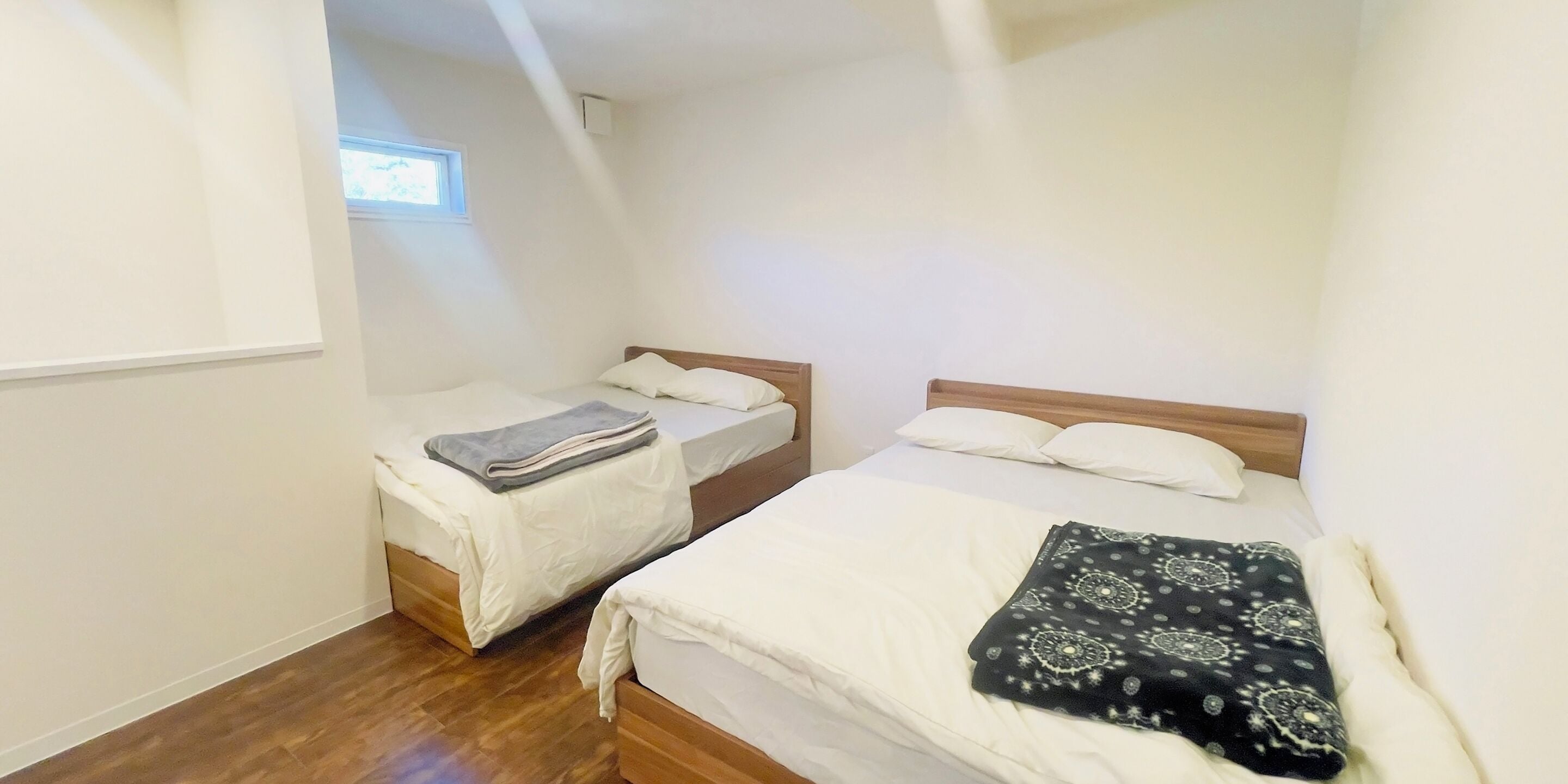 A small, clean room with two beds, a window, and a wooden floor.