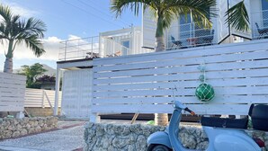 A white wooden fence with a stone wall in front, a scooter parked beside it, and a building with balconies in the background. - Vamos Villa Tokunoshima (Tokunoshima)