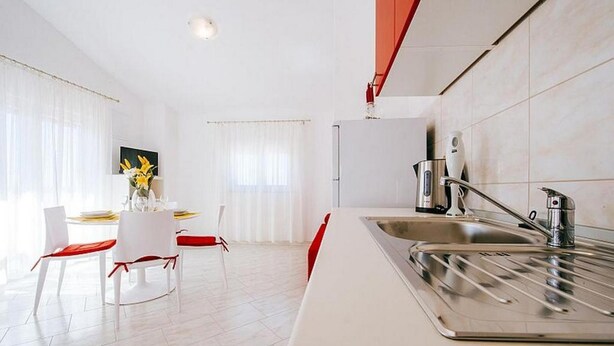 A modern kitchen with a stainless steel sink, white cabinetry, and a dining area with a table and chairs. - Apartments Perinic (Vrsi)
