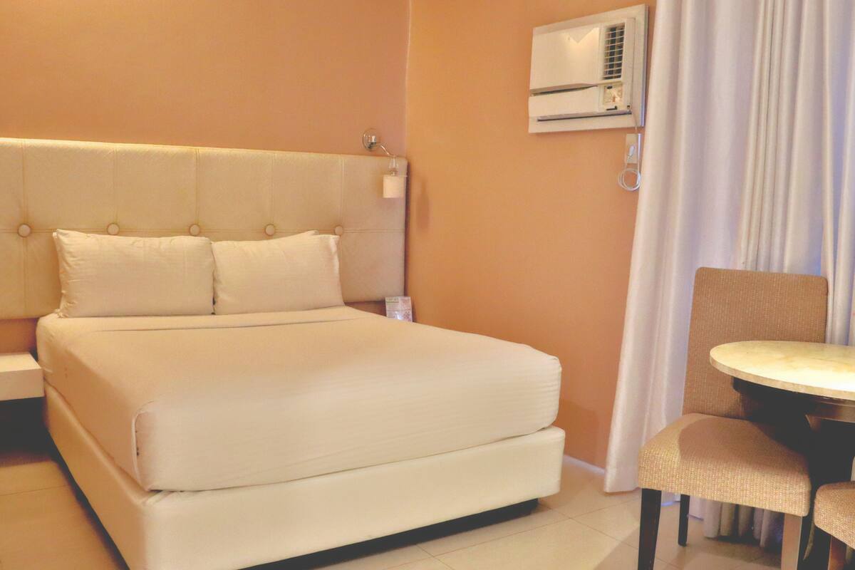 A hotel room with a large bed, a bedside table, a chair, and an air conditioning unit on the wall.